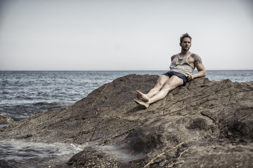 Handsome muscular man on the beach lying on rocks, looking at camera