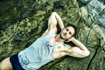Handsome muscular man on the beach lying on rocks, looking at camera