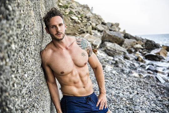 Handsome muscular shirtless man on the beach leaning on rock, looking at camera