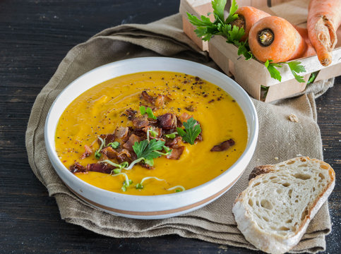 Roasted pumpkin and carrot soup with becon and pumpkin seeds on white wooden background.