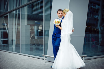 Groom in blue suit leans to a steel building while stading with
