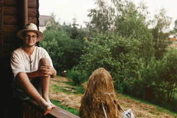 hipster man sitting on porch of wooden house  with field and hay