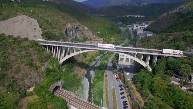 Lorry trucks move on road bridge in rock mountain up railway bridge 4k aerial drone view from above. Fly over arch bridge with vehicles move out of tunnel up river train railroad Italy France Europe