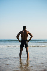 Fototapeta na wymiar Back view of black fit sportsman looking the sea for motivation before swimming. Male swimmer ready for outdoor training into the ocean.