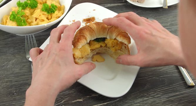 4K Fr&uuml;hst&uuml;ck Scene. Auf einem weissen eckigem Teller liegt ein Croissant belegt mit R&uuml;hrei und Speck. Eine Frau beisst gen&uuml;sslich in das H&ouml;rnchen.