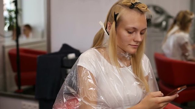 A blonde woman surfing the internet with her smartphone in hair studio. Slow motion