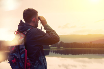 A traveler with a backpack looking through binoculars on the mountain and the lake. Vintage color,...