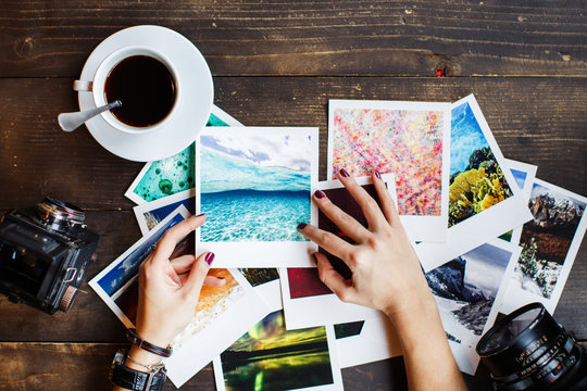 Women's Hands Holding Printed Photos. Dressed In Red Shirt, Lotus Tattoo On Arm. On Old Wooden Table Scattered Photos, Two Old Medium Format Film Camera, Glass Of Juice, Cup Of Coffee. Point Of View