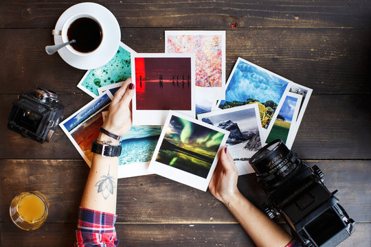 Women's hands holding printed photos. Dressed in red shirt, lotus tattoo on arm. On old wooden table scattered photos, two old medium format film camera, glass of juice, cup of coffee. Point of view