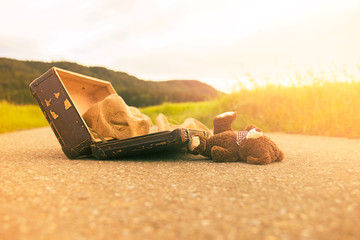 Abandoned, forsaken teddy bear in a vintage luggage suitcase on the asphalt road in the summer sun shine in the nature and beautiful light