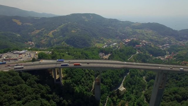 Bypass highway viaduct bridge above coastal sea town in Italy aerial 4k. Cars lorry truck cargo freight logistics. Vehicles move on massive bridge road above green valley top view from above by drone