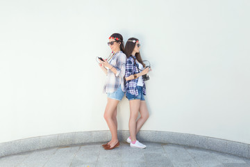 Two stylish young friends are standing back to back in front of the white wall and using phones. Girls are wearing matching outfits, plaid shirts and denim shorts.