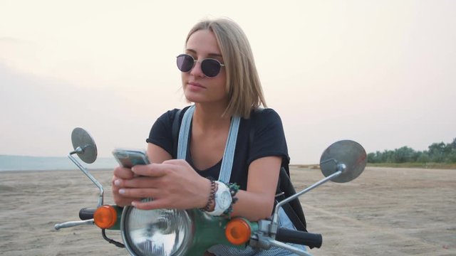 Young attractive female using smart phone while sitting on scooter on the beach