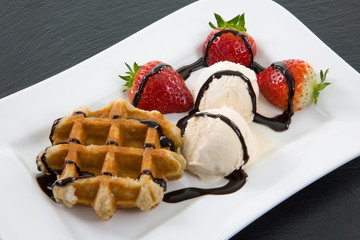Dessert aus Waffel, Eis und Erdbeeren, verziert mit Schokolade auf weissem Teller der auf einer Schieferplatte steht.