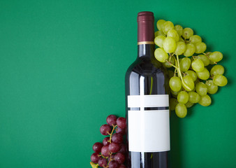 A full bottle of red wine with a blank label and bunches of grapes arranged on a green background...