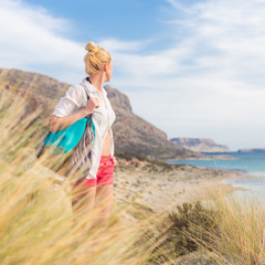 Relaxed woman on vacations in white loose shirt carrying beach bag and towel, enjoying beautiful...