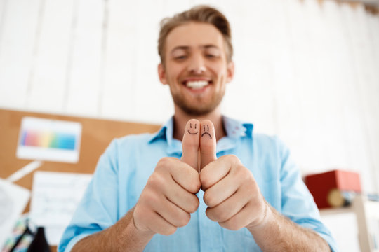 Young handsome smiling businessman showing thumbs up with funny faces drawings. Focus on hands. White modern office interior background.