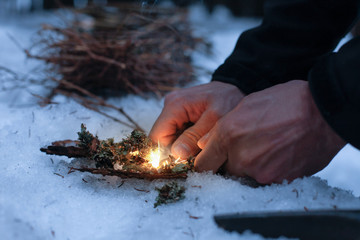 Man lighting a fire in a dark winter forest, preparing for an overnight sleep in nature, warming...