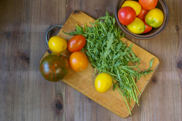 Fresh tomatoes and greens (arugula, parsley) on a cutting board on a wooden table. Near bowl with tomatoes