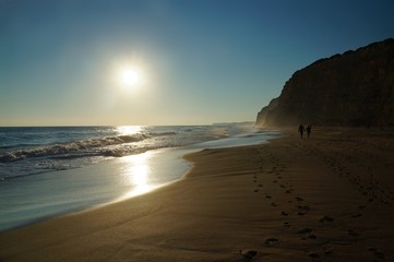 A couple strolls along the water's edge at high tide leaving two lines of foot prints leading down the beach. The sun is reflected on the water and on the wet sand. Algarve, Portugal.