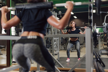 Young strong woman doing squats at the gym in front of a mirror with a barbell on her back
