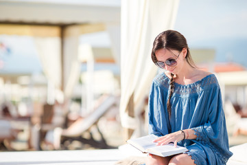 Young woman reading book during tropical beach vacation. Fashion girl read sitting in white sunbeds...