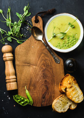 Fresh homemade pea cream soup in white bowl with bread over black backdrop with wooden serving board in center, top view, copy space, vertical composition