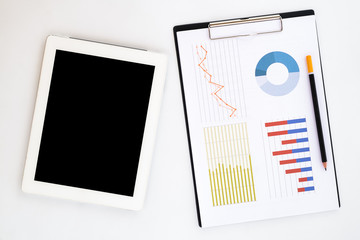 White office desk top with blank screen tablet, pencil and chart or graph over backboard. Top view with copy space