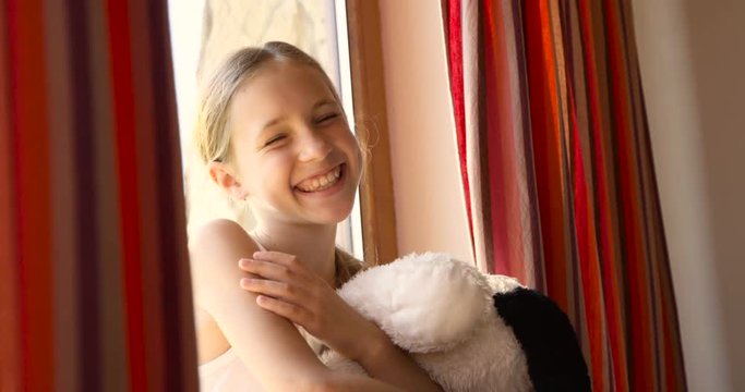 Cute beautiful little girl sitting on the windowsill with red-curtains and playing with her soft toy