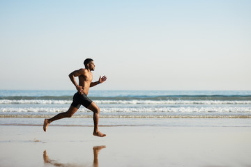 Black fit man running barefoot by the sea on the beach. Powerful runner training outdoor on summer. - Powered by Adobe