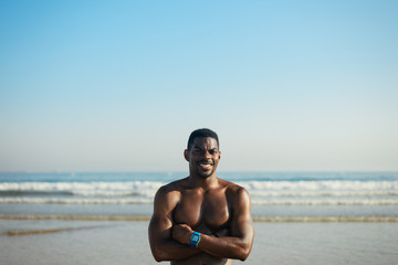 Fototapeta na wymiar Black cheerful man posing for portrait at the beach after swimming or running summer workout. Fit motivated athlete smiling and crossing arms towards sea background.