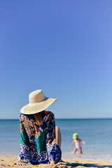 Fototapeta na wymiar Back view of young elegant female on sea beach sitting with hands up wearing dress and hat, sunny blue sky outdoors background