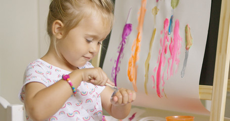 Little blond girl applying paint to the back of her hand with a smile using a paintbrush as she sits in front of an easel with a colorful abstract painting amusing herself at home.