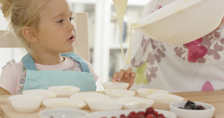 Happy little girl with empty muffin holders on table as parent picks up spoonful of batter from bowl
