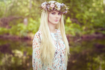 Beautiful blonde woman with flower wreath on her head. Beauty girl with flowers hairstyle. Girl in a summer forest. Fashion photo