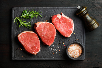 Raw beef Eye Round steaks with spices and rosemary on black slate stone board over dark wooden background, top view, horizontal composition