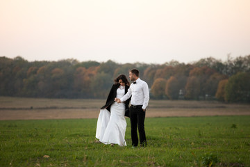 Bride in groom's jacket walks behind him on the green field