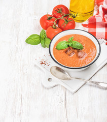 Cold gazpacho soup in bowl with ice, hot pepper and basil served with fresh tomotoes on ceramic board over white wooden background, top view, selective focus, copy space, vertical composition