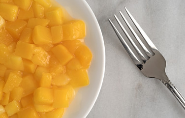 Diced mangoes on a white plate with fork atop a gray marble counter top.