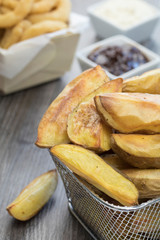 Oil free baked potato wedges in a mini wire basket on a wooden table with onion rings, barbeque sauce and mayonnaise in the background.