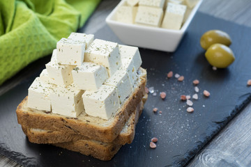 Tofu feta cheese with fresh oregano on a whole grain toast with linseed along with himalayan salt and two green olives on a black stone tray.