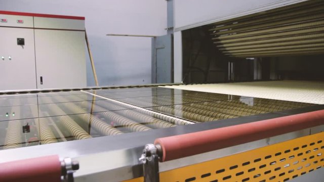 Widok na maszynę do hartowania szkła w fabryce. Elementy szklane leżą na rolkach i wjeżdżają do pieca. Hartowanie szkła