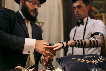 Groom stands straight while Jew puts on his hand black ribbon