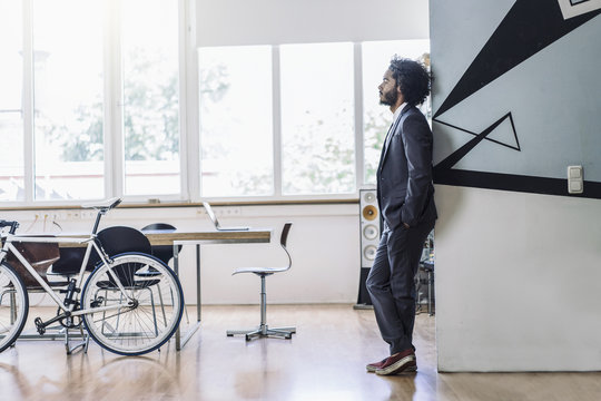 Young creative businessman standing in office, thinking with hands in pockets