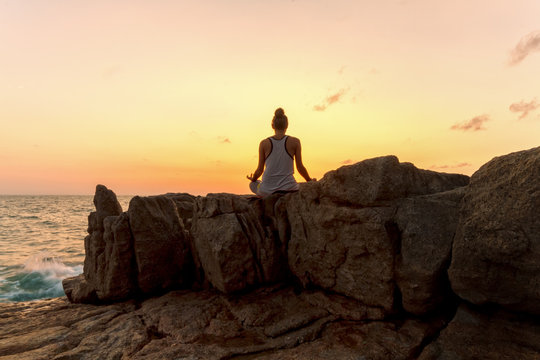 Young woman meditating on the rocks by the sea on sunrise background