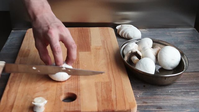Young man cooking. Cutting (slicing) white cultured mushrooms (chapignons) on kitchen board.