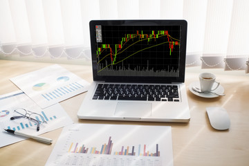 Stock graph on laptop screen with business report and cup of coffee on wooden table in home at morning time.