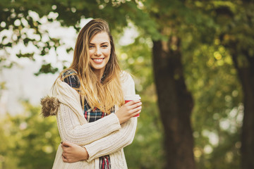 Beautiful young woman in sweater in park in autumn holding cup of takeaway coffee