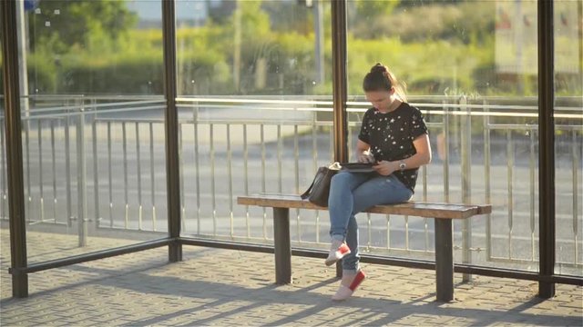 Young charming Caucasian female with trendy look use tablet while waiting transport on bus stop, woman sitting on a station against sunset background - Powered by Adobe