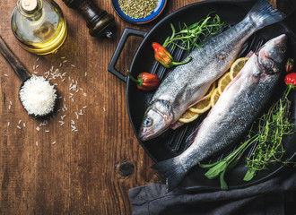 Ingredients for cookig healthy fish dinner. Raw uncooked seabass fish with rice, olive oil, lemon slices, herbs and spices on black grilling iron pan over rustic wooden background, top view, copy